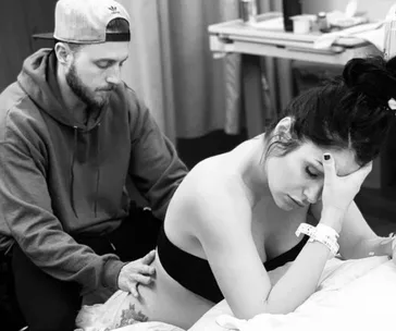 Man supporting woman in labor with back massage; she appears in pain, resting on bed.