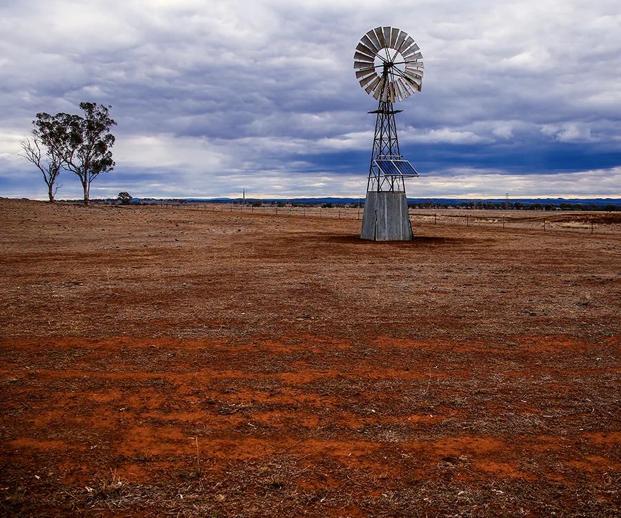 Windmill stands in an expansive, dry field under a cloudy sky, with distant trees and hills on the horizon.