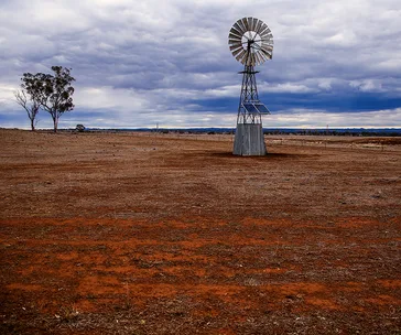 Windmill stands in an expansive, dry field under a cloudy sky, with distant trees and hills on the horizon.