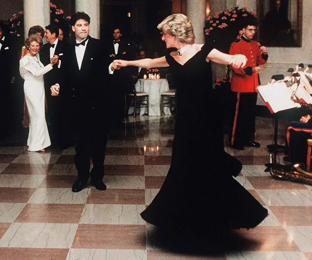 Princess Diana dances with a man in a tuxedo at a formal event, surrounded by guests and musicians.