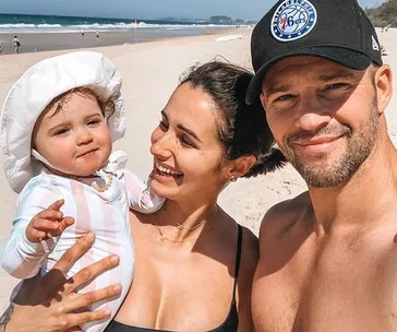 Family smiling at the beach; woman holding child with wide-brimmed hat, man in 76ers cap.