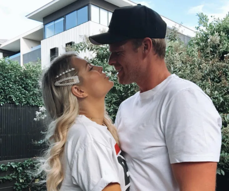 A couple smiling closely at each other in an outdoor setting, both wearing casual white shirts.