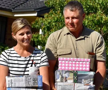 A smiling couple stands outdoors holding wrapped gifts against a sunny backdrop.