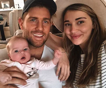 Family selfie of a smiling man, woman, and baby in a cozy living room setting.