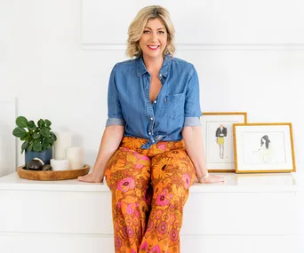 A woman in a denim shirt and vibrant floral pants sits on a white dresser, with framed art and candles nearby.