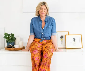 A woman in a denim shirt and vibrant floral pants sits on a white dresser, with framed art and candles nearby.