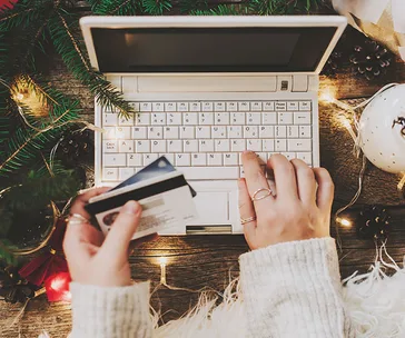 Hands holding credit cards near a laptop, surrounded by Christmas lights and decorations.