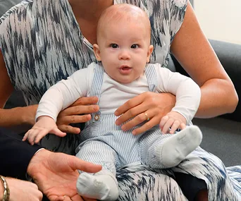Child sitting on an adult's lap, dressed in a white top and striped overalls, with attentive expression.