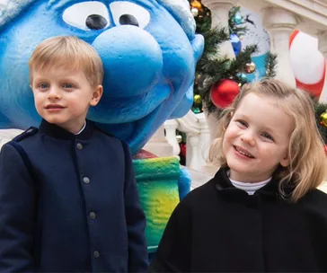Young boy and girl smile beside a large blue Smurf statue, with festive decorations in the background.