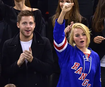 Two people cheer at a sports event; one wears a "Rangers" jersey, raising a fist, the other claps.