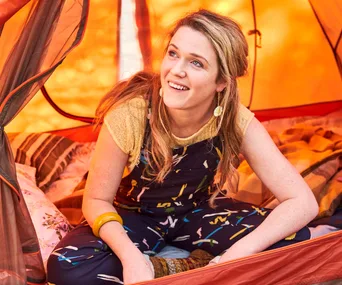 A woman smiling inside an orange tent, wearing colorful overalls and a yellow top.