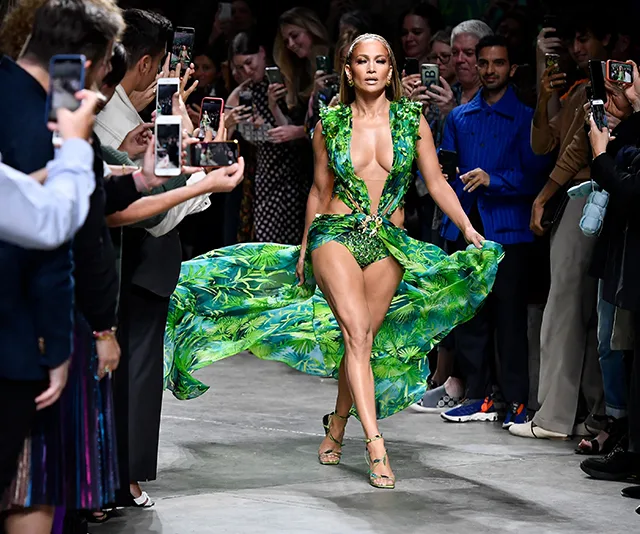Model in a green, tropical-print dress walks runway while onlookers take photos.