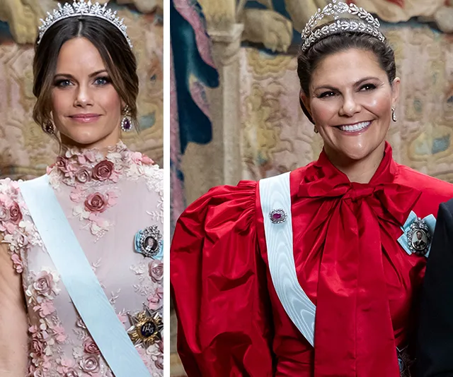 Two women in ornate dresses with tiaras, one in red and the other in floral pink, smiling at a formal event.