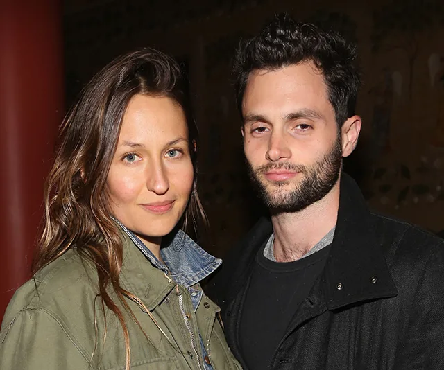 A couple posing together, both with casual attire and neutral expressions, in a warmly lit indoor setting.