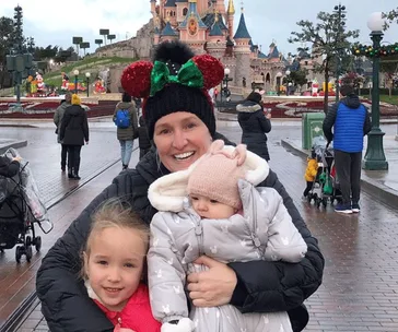 Family enjoying Disneyland, mother in Minnie Mouse ears holds two kids in front of Sleeping Beauty Castle.