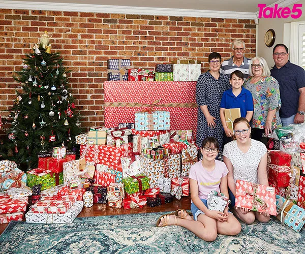Family posing with numerous wrapped presents by a Christmas tree inside a living room, smiling for the camera.