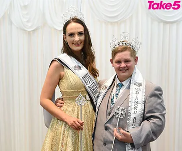 Two pageant winners wearing crowns and sashes, smiling, standing in front of a white curtain backdrop.