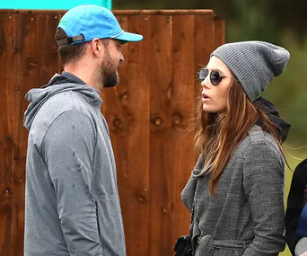 Two people outdoors, one wearing a blue cap and gray hoodie, the other in a gray beanie and coat, talking.