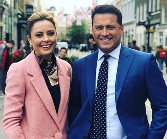 Two smiling people in formal attire standing on a street with Union Jack flags in the background.