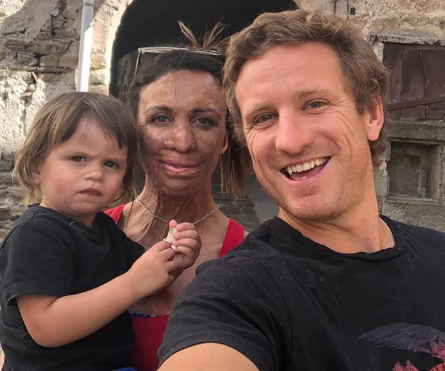 A family poses for a selfie, with a mother in the center holding a young child, and a smiling man beside them against a stone backdrop.