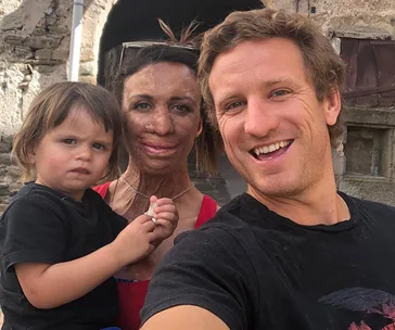 A family poses for a selfie, with a mother in the center holding a young child, and a smiling man beside them against a stone backdrop.