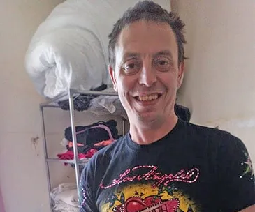 Man smiling in a room with shelves of folded clothes and a rolled-up blanket in the background.