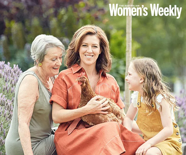 Three women outdoors, sitting, holding a chicken. Smiling together, surrounded by greenery.