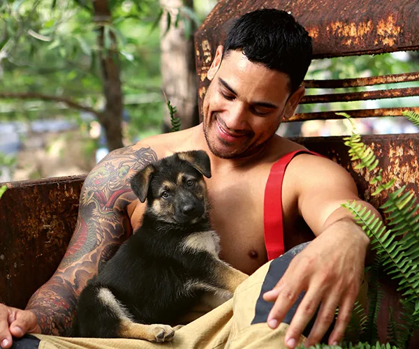 Firefighter with tattoos smiling at a German Shepherd puppy on his lap, surrounded by greenery and rusted metal backdrop.