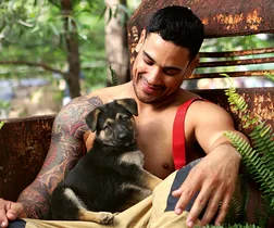 Firefighter with tattoos smiling at a German Shepherd puppy on his lap, surrounded by greenery and rusted metal backdrop.