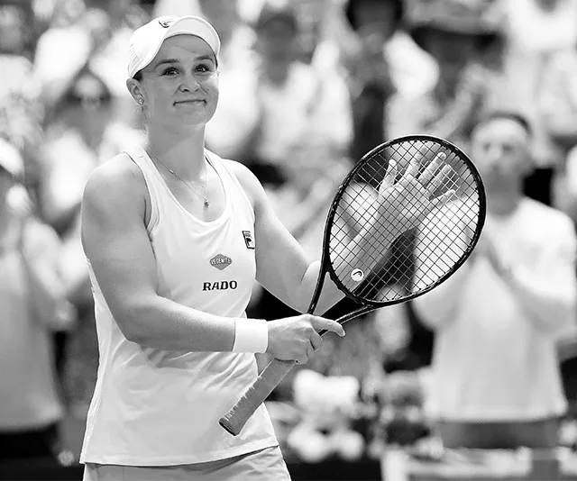 Tennis player holding a racket and waving to the crowd, wearing a cap and sleeveless shirt, on a court.