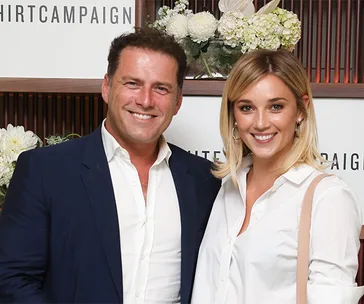 A smiling man in a suit and a woman in a white shirt at an event with floral decor in the background.