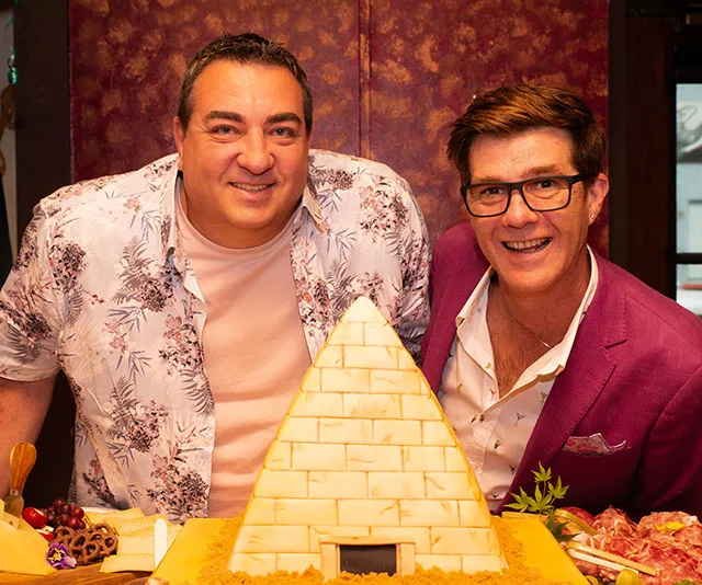 Two men smiling beside a pyramid-shaped cake; one wears a floral shirt, the other a pink blazer.