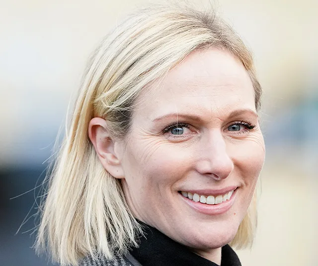 Blonde woman smiling, wearing a black outfit with a blurred, neutral background.