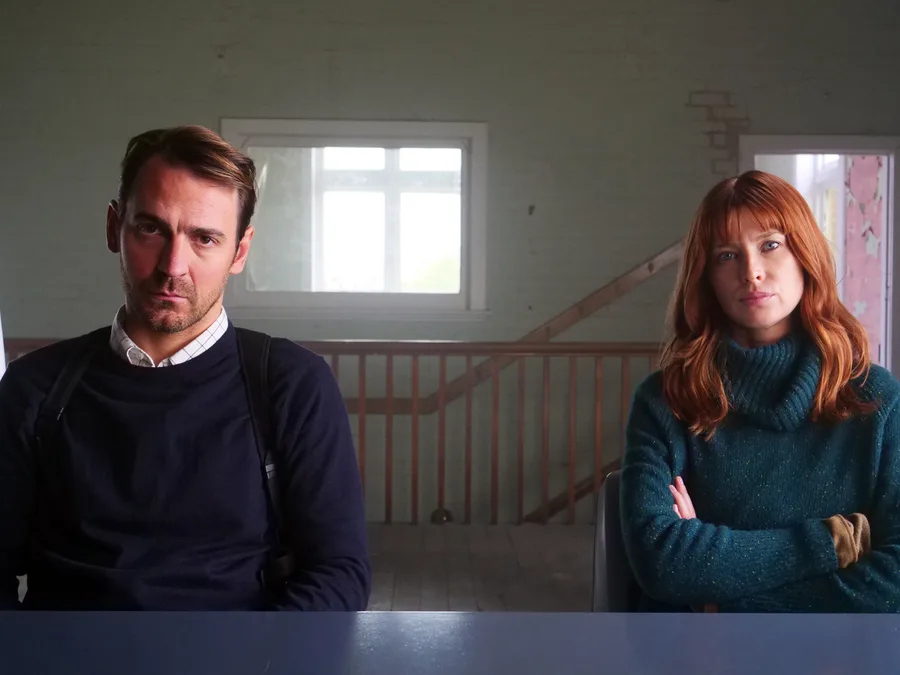 Two people sitting at a table in a dimly lit room, both looking serious and thoughtful.