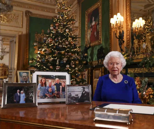 Queen Elizabeth II sits at a desk with family photos and Christmas decor in an ornate room.