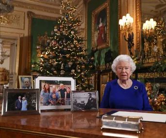 Queen Elizabeth II sits at a desk with family photos and Christmas decor in an ornate room.