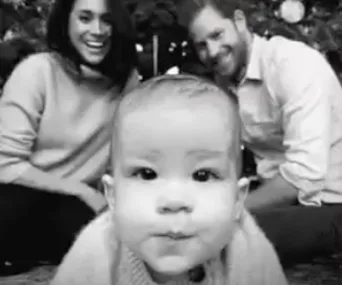 A baby with two adults smiling in the background, in front of a Christmas tree.