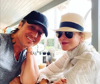 A couple smiling at a cafe, with one wearing a black cap and the other in a white hat and sunglasses.