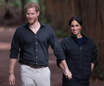 A couple walks hand in hand in a forest, both smiling and casually dressed.