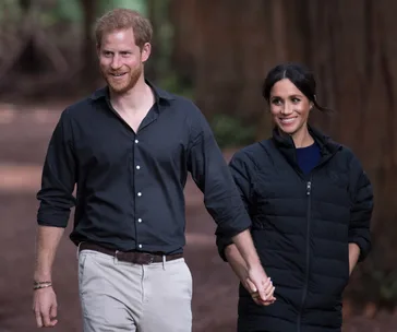 A couple walks hand in hand in a forest, both smiling and casually dressed.