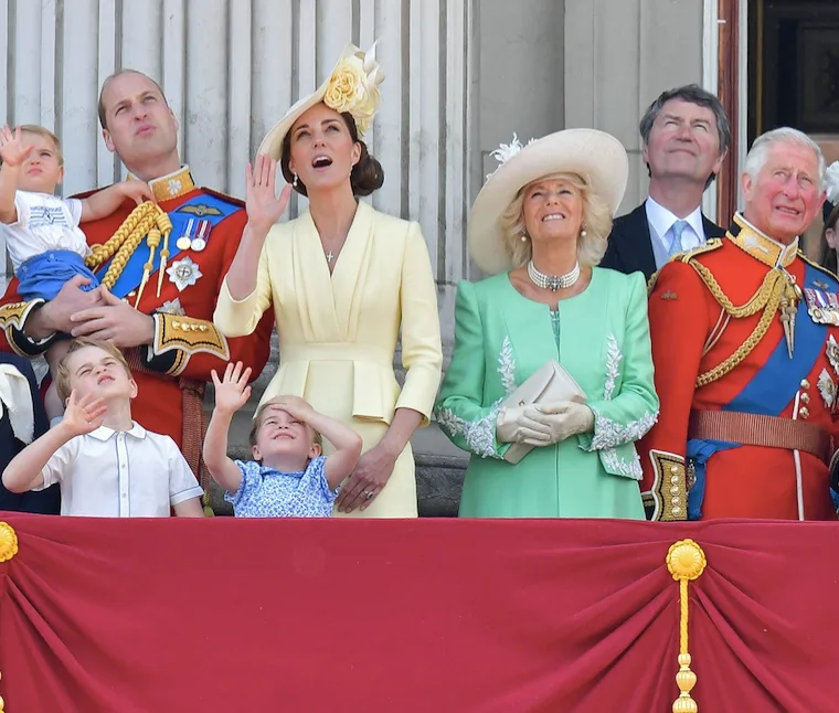 Royal family members on a balcony during an event, waving and looking up while dressed in formal attire.