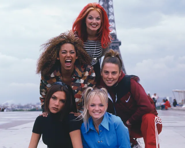 Five women posing and smiling near the Eiffel Tower with a cloudy sky in the background.