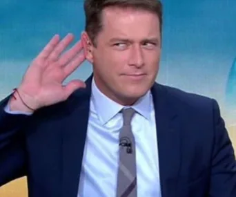 Man in a suit raises hand to ear, making a listening gesture, with a playful expression on a televised show set.
