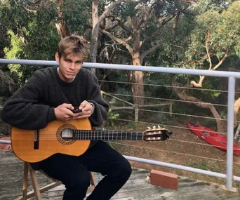 Person sitting outdoors with a guitar, holding a phone, surrounded by trees.