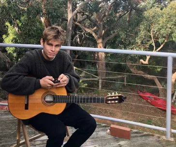 Person sitting outdoors with a guitar, holding a phone, surrounded by trees.