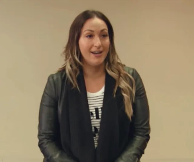 A woman with long hair, wearing a leather jacket and striped shirt, stands speaking in front of a plain background.