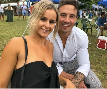 Smiling couple poses outdoors at a casual event with people and tents in background.