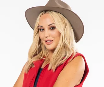 A woman with long blonde hair in a red vest and brown hat, smiling, poses against a white background.