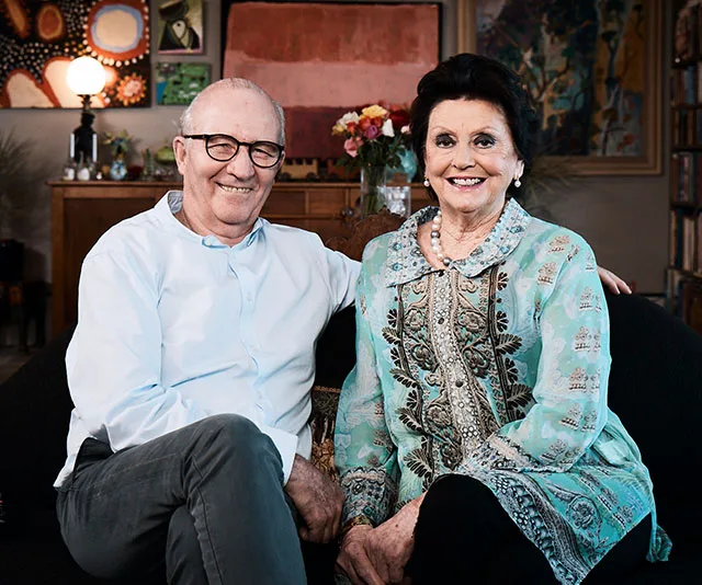 Elderly couple sitting on a couch, smiling warmly, in a cozy, art-filled living room.