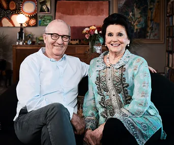 Elderly couple sitting on a couch, smiling warmly, in a cozy, art-filled living room.